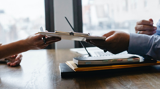 two hands handing off a clipboard to each other at a desk